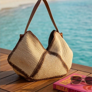 Bolsa de praia de palha Lívia sobre deck de madeira com óculos de sol e livro, mar ao fundo